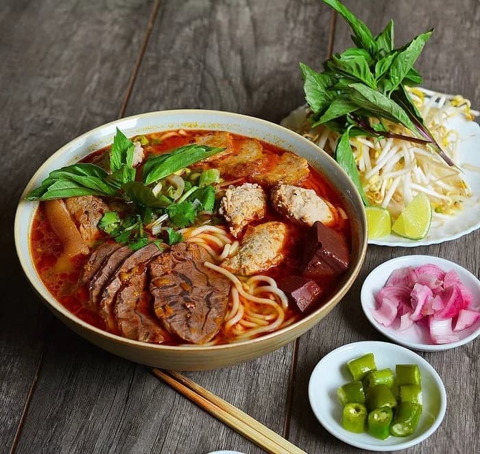 A bowl of bun bo Hue: round rice noodles and sliced beef in a chilli-red broth, topped with crab and pork balls and fresh herbs, with bean sprouts, lime, pickled onion and green chilli alongside