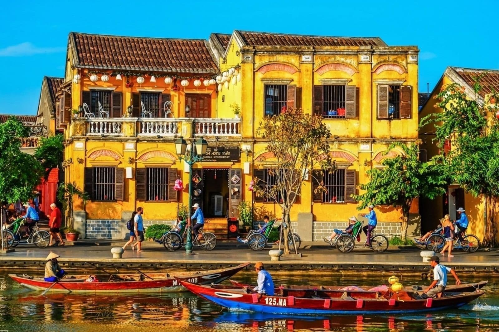 Two-storey yellow shophouses with brown shutters and strings of white lanterns along a riverside street, with cyclos and walkers on the promenade and wooden boats on the water in front