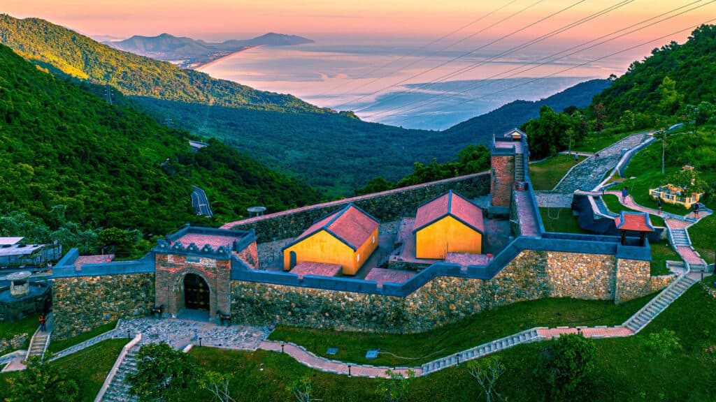 A restored stone fort with a gatehouse and two yellow buildings on a forested ridge under a pink sky, overlooking a bay and a long sandy spit